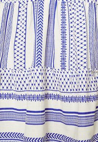 Vitt tyg med blå geometriska mönster, inklusive ränder, hexagoner och zickzack-mönster, med en volangkant.