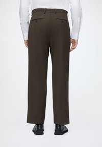 Homme portant un pantalon marron foncé sur mesure, une chemise blanche et des chaussures de ville noires, debout de dos sur un fond clair uni.