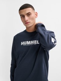 Junger Mann, der einen marineblauen Sweatshirt mit dem Schriftzug "HUMMEL SPORTSWEAR SINCE 1923" trägt und seinen Hals berührt, vor einem einfarbigen hellen Hintergrund.