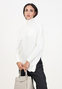 Maglione a collo alto bianco a costine con spacchi laterali, abbinato a una borsa grigia chiara. La texture mostra linee verticali prominenti e un materiale accogliente.