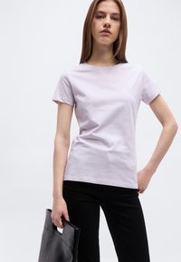 Γυναίκα που φοράει ανοιχτό μωβ κοντομάνικη μπλούζα και μαύρο παντελόνι, κρατώντας μια μαύρη τσάντα ώμου μπροστά σε απλό φωτεινό φόντο.