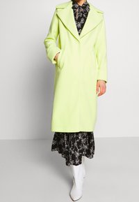 Neon green wool blend coat with a lapel collar, side pockets, and a straight silhouette over a black floral dress and white ankle boots.