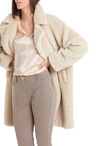 Manteau beige duveteux à large col, associé à un haut en satin clair et un pantalon à carreaux taille haute avec poches latérales et décorations.