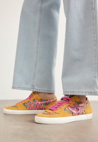 Baskets en daim jaune avec un tissu coloré et à motifs et des lacets roses. Elles présentent une semelle en caoutchouc blanche et un design texturé sur le dessus.