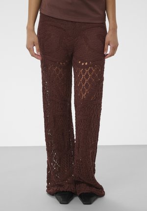 Pantalon marron en crochet avec des motifs ajourés complexes, porté avec des chaussures noires pointues et un haut marron assorti.