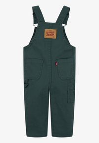 Grüne Baumwoll-Overalls mit verstellbaren Trägern, einer vorderen aufgesetzten Tasche und seitlichen Cargo-Taschen für zusätzlichen Stauraum. Robustes Material.