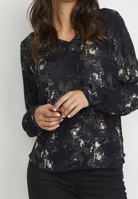 Femme portant un haut noir à col en V avec des motifs floraux abstraits blancs et verts, se tenant les mains jointes devant elle, accompagnée de pantalons noirs.