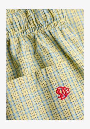 Geel-blauw geruit stof met geplooid elastiek en een rood geborduurd "S. Co"-logo op een zak.