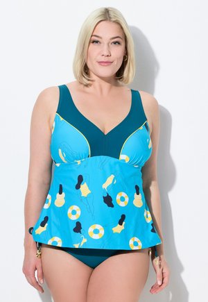 Frau trägt ein blaues Tankini mit Aufdruck von Schwimmern und Schwimmringen, steht vor weißem Hintergrund und schaut nach vorne.