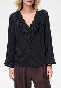 Kobieta w czarnej bluzce z długim rękawem i falbankowym dekoltem w kształcie litery V oraz brązowych spodniach, prezentująca tatuaż na lewej dłoni.