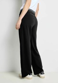 Svarta vida byxor med en slät textur och en åtsittande midja, med en bakficka, ihopparade med vita sneakers.