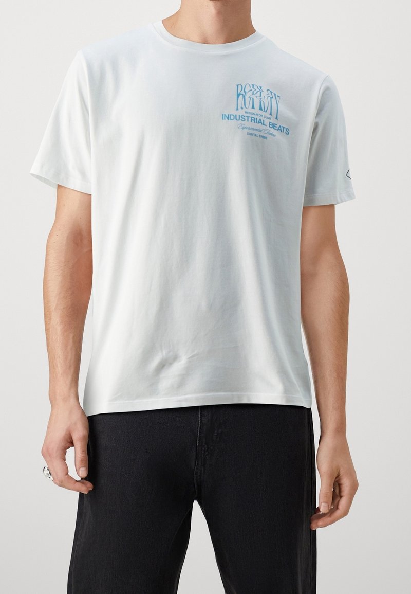 T-shirt en coton blanc à manches courtes, col rond, avec un imprimé graphique en texte bleu sur la poitrine et un petit logo sur la manche.