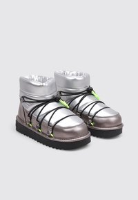 Bottes argentées et grises avec une tige rembourrée, des lacets noirs et des accents vert vif. Elles présentent une semelle noire épaisse et un éclat métallique.