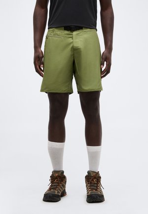 Homme portant un short vert olive, des chaussettes blanches montantes et des chaussures de randonnée marron, debout devant un fond uni.