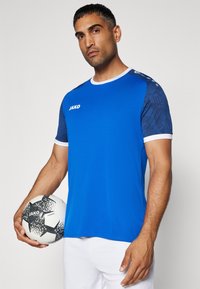 Maglietta atletica blu con dettagli bianchi e maniche a fantasia, abbinata a un pallone da calcio bianco e nero. Presenta dettagli di marca.