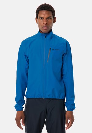 Uomo che indossa una giacca blu brillante con cerniera e tasca sul petto sopra pantaloni neri, in piedi davanti a uno sfondo chiaro e uniforme.