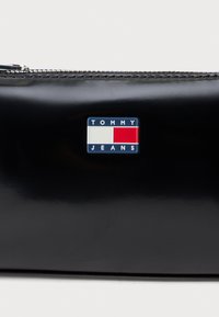 Pochette en cuir noir brillant avec fermeture éclair argentée et badge logo rectangulaire en blanc, rouge et bleu indiquant "Tommy Jeans" au centre sur le devant.