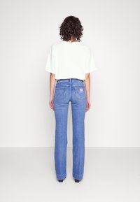 Jean flare bleu à taille haute, avec un détail de couture sur les poches arrière et un t-shirt blanc, vu de l'arrière sur fond clair.