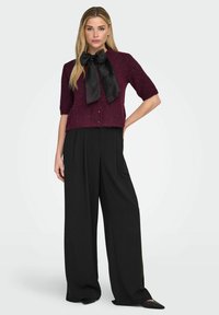 Cardigan lavorato a maglia bordeaux a maniche corte con fiocco in raso nero, bottoni sul davanti, abbinato a pantaloni neri a gamba larga e vita alta.