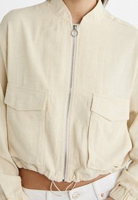 Chaqueta corta color crema hecha de tela texturizada, con cuello alto, cierre de cremallera y dos bolsillos frontales con dobladillo elástico.