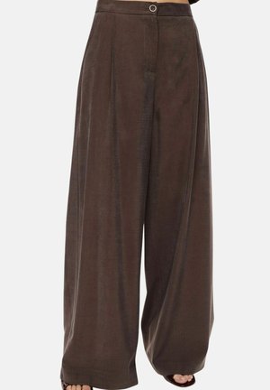 Pantaloni marroni a gamba larga con vita alta, chiusura frontale con bottone e cerniera, indossati con sandali scuri aperti, mostrati dalla vita ai piedi.