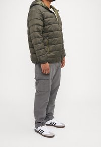 Olivgrön vadderad jacka med huva och horisontell sömnad. Kombinerad med grå cargobyxor och vita sneakers med svarta ränder.