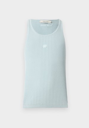 Jeune homme debout, main dans la poche, portant un haut côtelé sans manches bleu clair, un pantalon crème et des mocassins marron et blanc sur fond blanc.