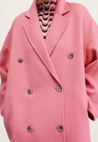 Růžový vlněný kabát s dvojitým zapínáním, velkými knoflíky, širokými klopami a spadenými rameny, přehozený přes barevně pruhovaný rolák.