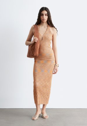 Femme aux longs cheveux foncés porte une robe à motif orange avec un col licou, porte un sac à épaule marron et des sandales beige à brides.