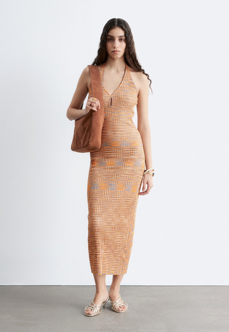 Femme aux longs cheveux foncés porte une robe à motif orange avec un col licou, porte un sac à épaule marron et des sandales beige à brides.