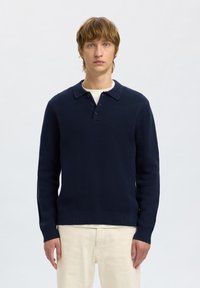 Suéter de punto azul marino con cuello en forma de camisa, tres botones y mangas largas, combinado con pantalones de color claro.