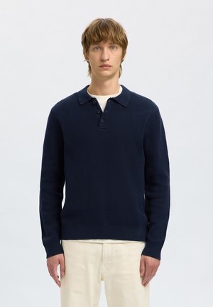 Navyblauwe gebreide pullover trui met een kraag-halslijn, drie knopen en lange mouwen, gecombineerd met lichte broek.