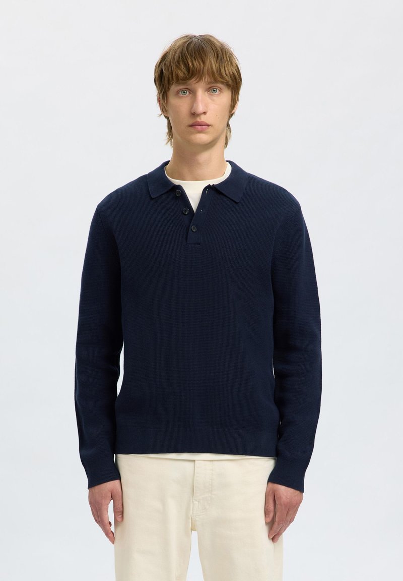 Suéter de punto azul marino con cuello en forma de camisa, tres botones y mangas largas, combinado con pantalones de color claro.