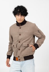 Mann mit lockigem schwarzen Haar, der eine braune, geknöpfte Wolljacke mit vorderen Taschen und schwarz gerippten Manschetten und Kragen trägt, posiert mit einer Hand in der Tasche.