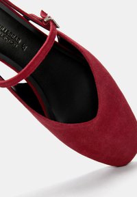 Rote Wildleder-Pumps mit offenem Zehenbereich und Knöchelriemen. Schwarze Innensohle mit Branding. Glatte Textur und geschwungene Form.