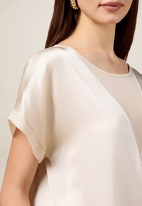 Beige zijden blouse met een V-hals en korte mouwen. Geen structuur met een subtiele glans, met een gelaagd ontwerp in bijpassende tinten.