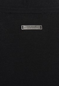 Zwarte gebreide trui stof met een zilveren rechthoekig label met "ESPRIT" erin geperst, gepositioneerd nabij de halslijn.