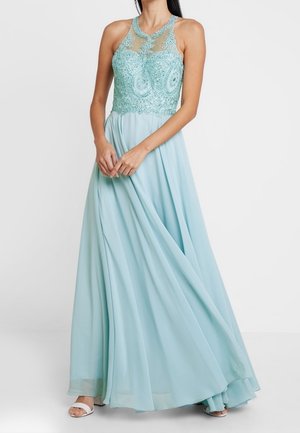 Femme portant une robe longue bleu clair avec des détails en dentelle et perles sur un col halter transparent, tenant la jupe fluide des deux mains.