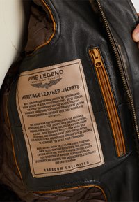 Unutrašnjost crne kožne jakne sa smeškom oznakom "Heritage Leather Jackets." Sadrži bež šavove i džep sa rajsferšlusom.