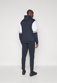 Μαύρο φούτερ με κουκούλα και λευκά μανίκια με ριγωτές λεπτομέρειες, συνδυασμένο με μαύρο παντελόνι και μαύρα παπούτσια. Το ύφασμα φαίνεται λείο και αθλητικό.