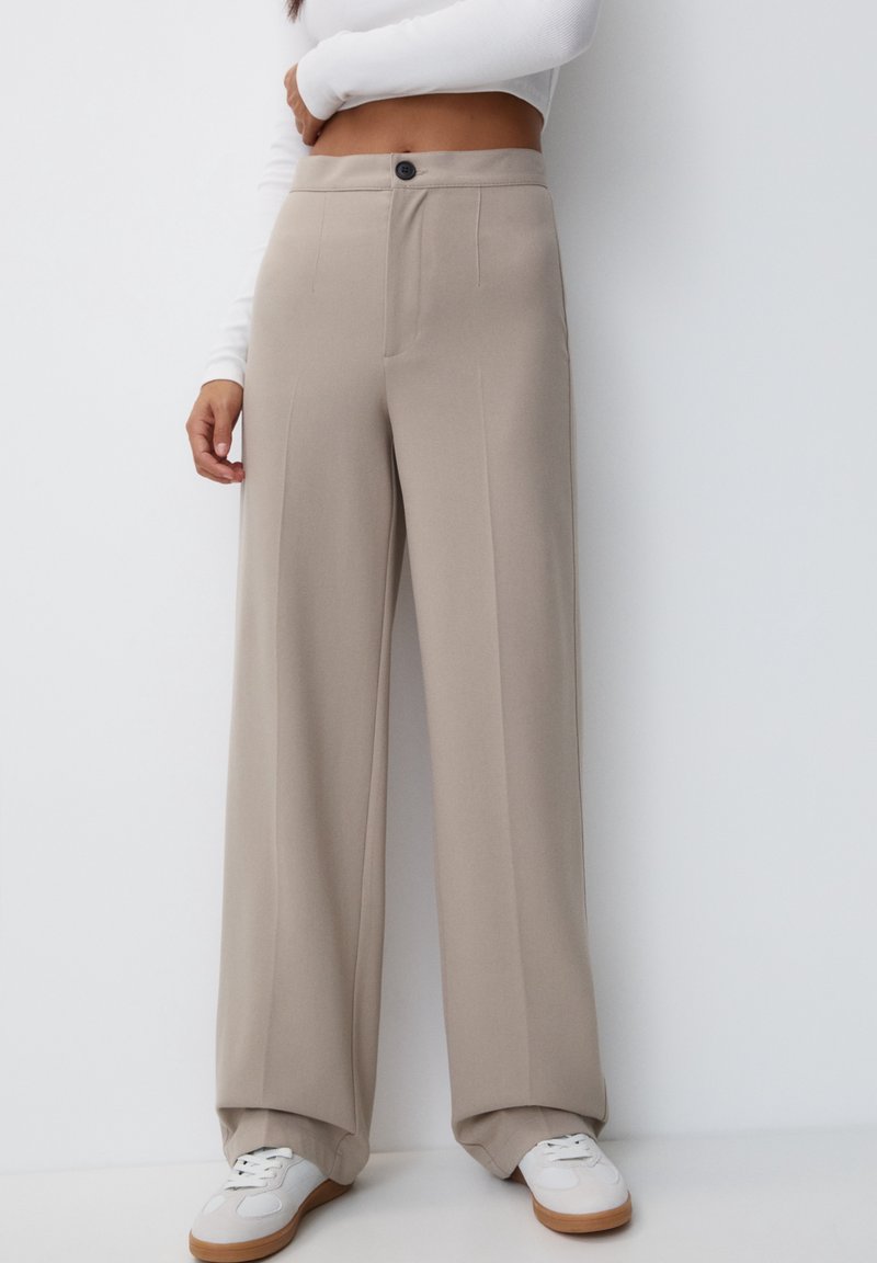 Femme portant un pantalon large beige, un top court blanc à manches longues et des baskets blanches, debout sur un fond blanc.