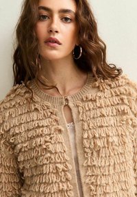 Cardigan beige lavorato a maglia con un motivo testurizzato a anelli, scollatura rotonda e un piccolo apertura a forma di chiave nella parte anteriore, abbinato a un top leggero sottostante.