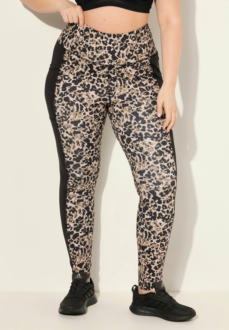 Leopard-Print Leggings in Beige, Schwarz und Braun. Hoher Taillenbund mit kontrastierenden schwarzen Seitenbahnen. Weiches, elastisches Material. Schwarze Sneaker.