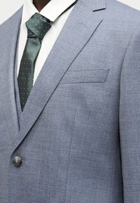 Άνδρας που φοράει ανοιχτό μπλε σακάκι με υφή πάνω από λευκό πουκάμισο με σκούρα πράσινη γραβάτα διακοσμημένη με μικρές λευκές κουκκίδες, κουμπωμένη εμπρός.