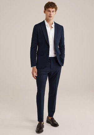 Traje azul marino de corte ajustado, chaqueta de un solo botón, solapas con muesca y camisa blanca. Mocasines de cuero negro completan el look.