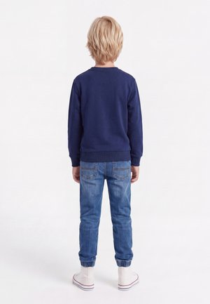 Niño rubio de pie de espaldas, vistiendo una sudadera azul marino, pantalones vaqueros deportivos azules y zapatillas altas blancas sobre un fondo blanco.