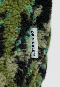 Gestructureerde groene en zwarte stof met een camouflagepatroon. Een genaaid label met de tekst "ORIGINALS™" in het wit op een zwarte achtergrond.