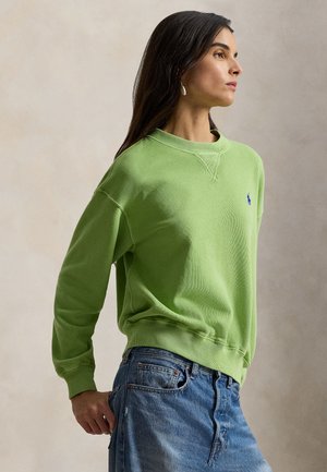 Kvinde med langt mørkt hår iført en lysegrøn sweatshirt og blå jeans, stående i profil mod en neutral baggrund.