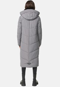 Long gray puffer coat with a hood, featuring chevron stitching, a mid-calf length, and a branded tag near the bottom. Worn with black pants and boots.