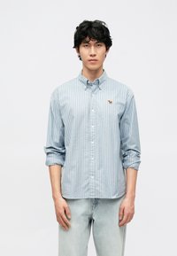 Φωτεινό γαλάζιο πουκάμισο με ρίγες και κουμπιά, κατασκευασμένο από βαμβάκι, με μικρή κεντημένη λεπτομέρεια. Μανίκια γυρισμένα, σε συνδυασμό με ανοιχτόχρωμο τζιν.